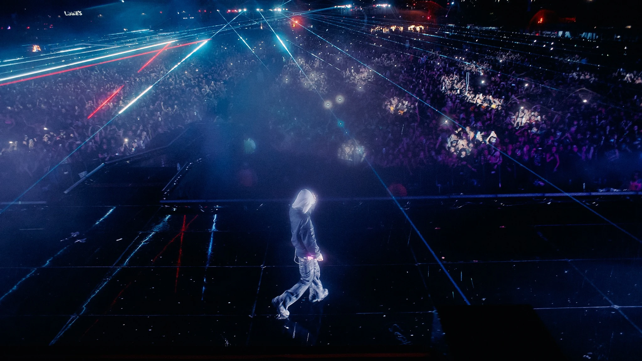 A hooded performer walking across the stage beneath lasers