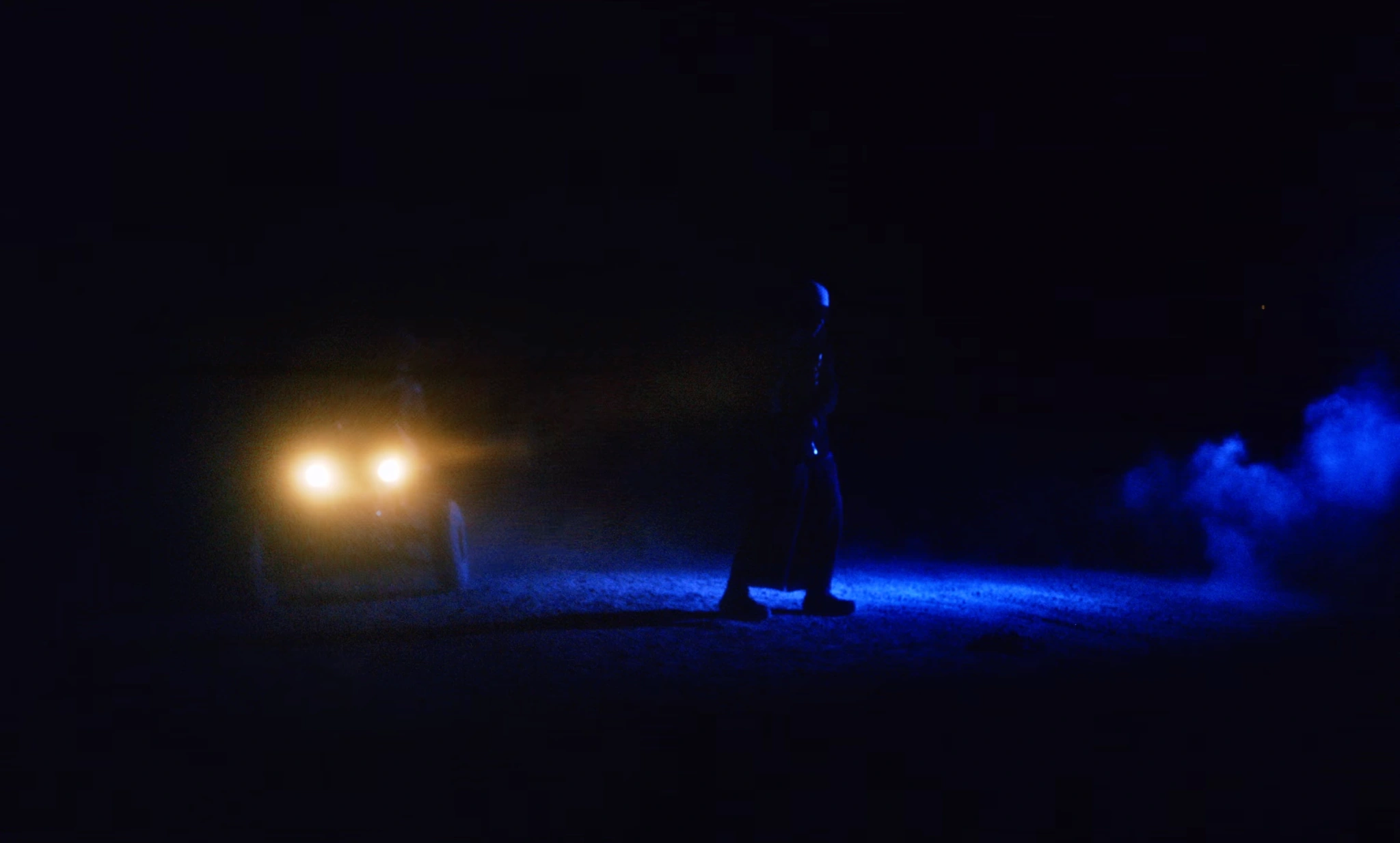 A figure standing between headlights and blue smoke at night