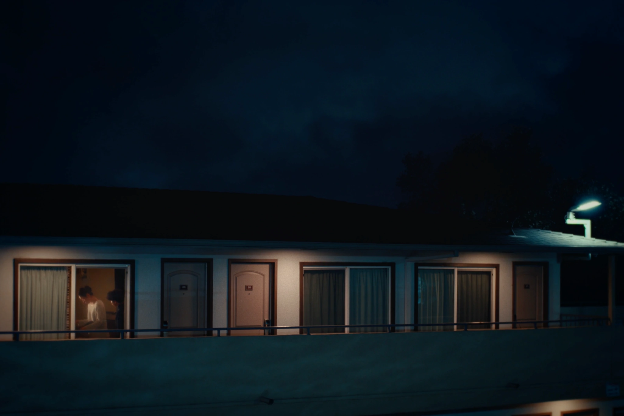 Night exterior of an apartment walkway from Sweet Boy, with two figures visible through a window